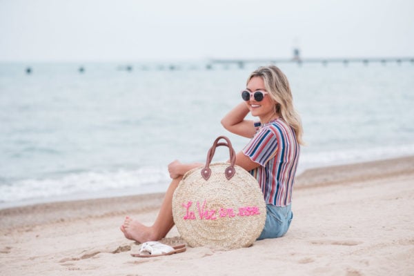 Chicago fashion blogger Jessica Sturdy wearing pink sunglasses, Bershka candy striped shirt, Cotton On high-rise shorts with Soludos slide sandals and a White Elephant Designs embroidered straw tote with "La Vie en Rose" collaboration with Jessica Sturdy at Lake Michigan in Chicago.