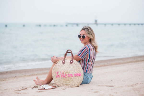 Chicago fashion blogger Jessica Sturdy wearing pink sunglasses, Bershka candy striped shirt, Cotton On high-rise shorts with Soludos slide sandals and a White Elephant Designs embroidered straw tote with "La Vie en Rose" collaboration with Jessica Sturdy at Lake Michigan in Chicago.