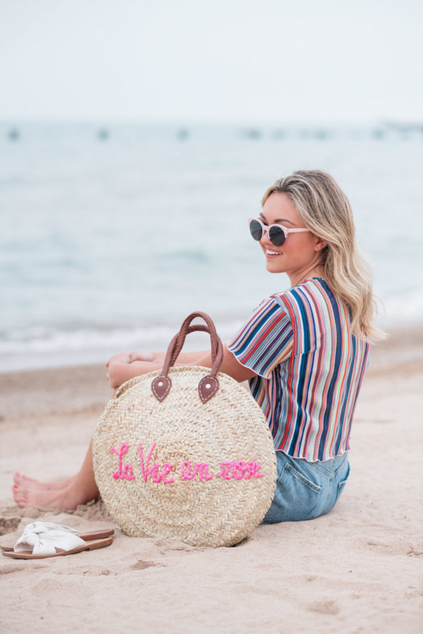 Chicago fashion blogger Jessica Sturdy wearing pink sunglasses, Bershka candy striped shirt, Cotton On high-rise shorts with Soludos slide sandals and a White Elephant Designs embroidered straw tote with "La Vie en Rose" collaboration with Jessica Sturdy at Lake Michigan in Chicago.