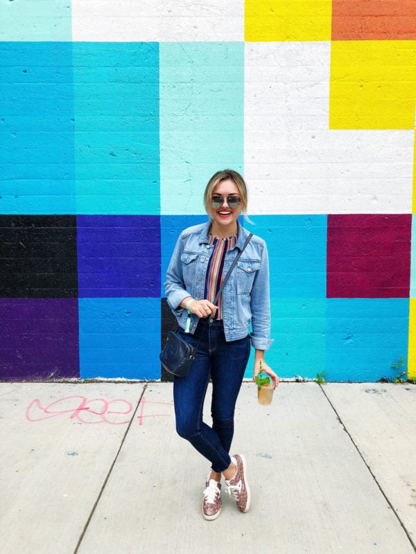 Chicago lifestyle blogger Jessica Sturdy wearing a denim jacket with blue jeans, glitter sneakers, Celine sunglasses, and a Gucci crossbody bag at a concert in Chicago in front of a colorful mural wall.