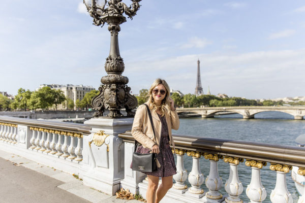 Jessica Sturdy (blogger behind Bows & Sequins) in Paris on a gorgeous, sunny September Day. She is on one of the prettiest, gilded bridges in Paris, with magnificent views of the Eiffel Tower. She's wearing a Sandro leather jacket, a flirty floral dress, and a Lancel Pia bag.