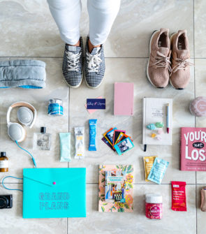 Fashion and travel blogger Jessica Sturdy of Bows & Sequins shares a packing flat lay photo for what she brought along on Remote Year. She's wearing gingham sneakers and standing in the photo with white jeans surrounded by Bose headphones, books, vitamins, RXBars, polaroid photos, and other wellness items that focus on fitness and mindfulness.