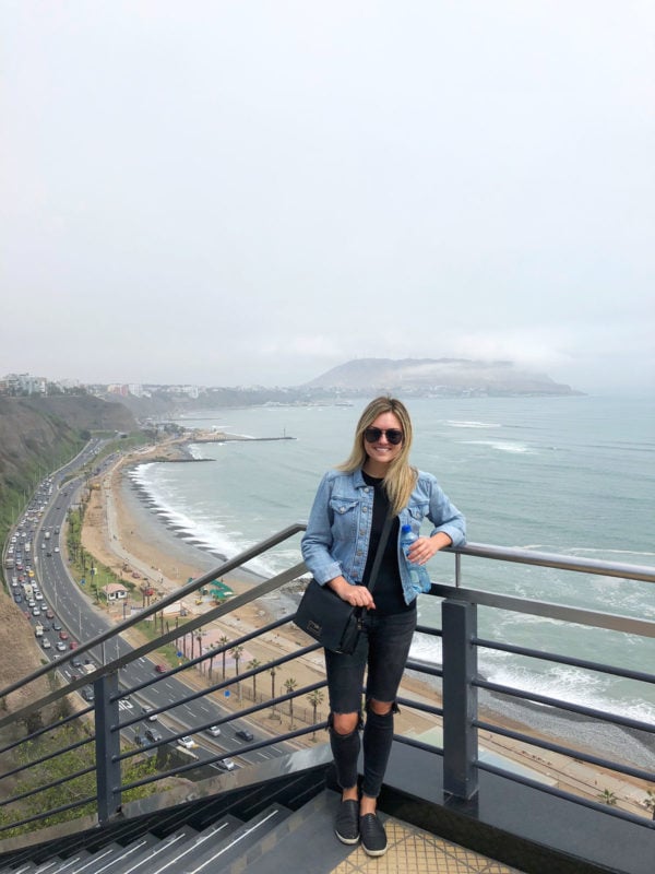 Remote Year blogger Jessica Sturdy of Bows & Sequins at Larcomar in Lima Peru with gorgeous views of the Lima cliffside and Pacific Ocean. Jessica is wearing black jeans and a blue jean jacket.