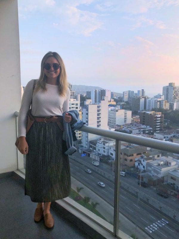 Remote Year blogger Bows & Sequins wearing a pleated green skirt and cream sweater with leather accessories on her apartment balcony in Miraflores, Lima, Peru.