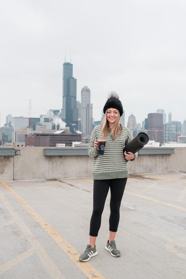 Fitness influencer Jessica Sturdy of Bows & Sequins wearing a mink beanie, American Eagle striped henley sweater, Lululemon leggings, and Adidas Flashback Sneakers with a Dunkin Donuts Energy Cold Brew and a yoga mat at the Core Power Yoga in the South Loop of Chicago with a view of the skyline and Willis Tower.