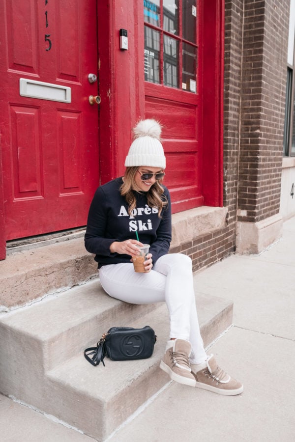 Chicago health and wellness influencer Jessica Sturdy sitting on a step in front of a red door wearing white jeans, shearling sneakers, and a pom-pom beanie.