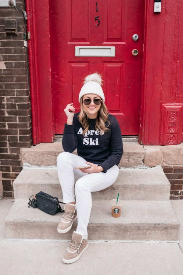 Everyday lifestyle blogger Jessica Sturdy sitting on a step in front of a red door in Chicago wearing a casual weekend outfit. She's styling a Yves Salomon beanie with Gucci aviators, an Apres Ski sweater, white jeans from Old Navy, a Gucci crossbody bag, and shearling-lined sneakers from Marc Fisher via Nordstrom.