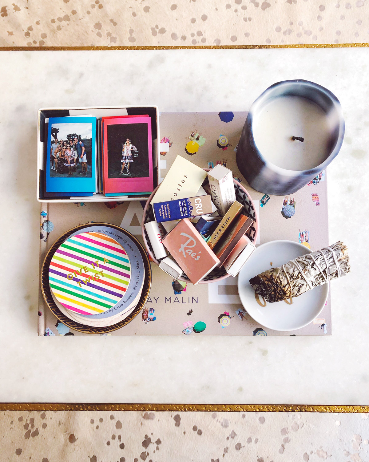 Jessica Sturdy styles the white marble coffee table in her Gold Coast apartment in Chicago. A blue and white candle, a white sage smudge stick, and colorfully striped Kate Spade Coasters are atop a Gray Malin Beaches book, along with a tray full of mini Fujifilm Instax polaroid pictures and a bowl full of matchbooks collected from her travels.