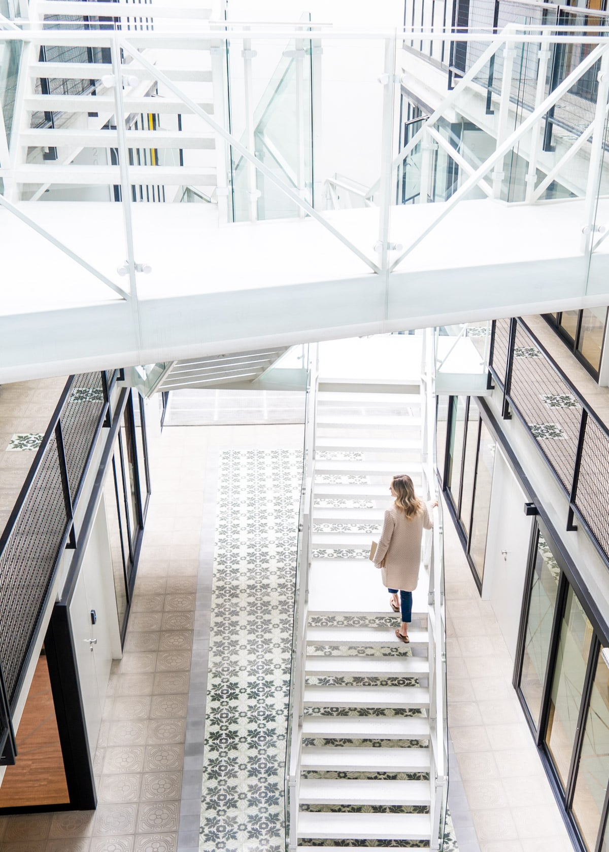 Jessica Sturdy on a white twisted staircase at Comunal Co-Working in Lima, Peru. She is carrying a laptop and wearing a long cashmere cardigan with cropped jeans and leather slip-on loafers.