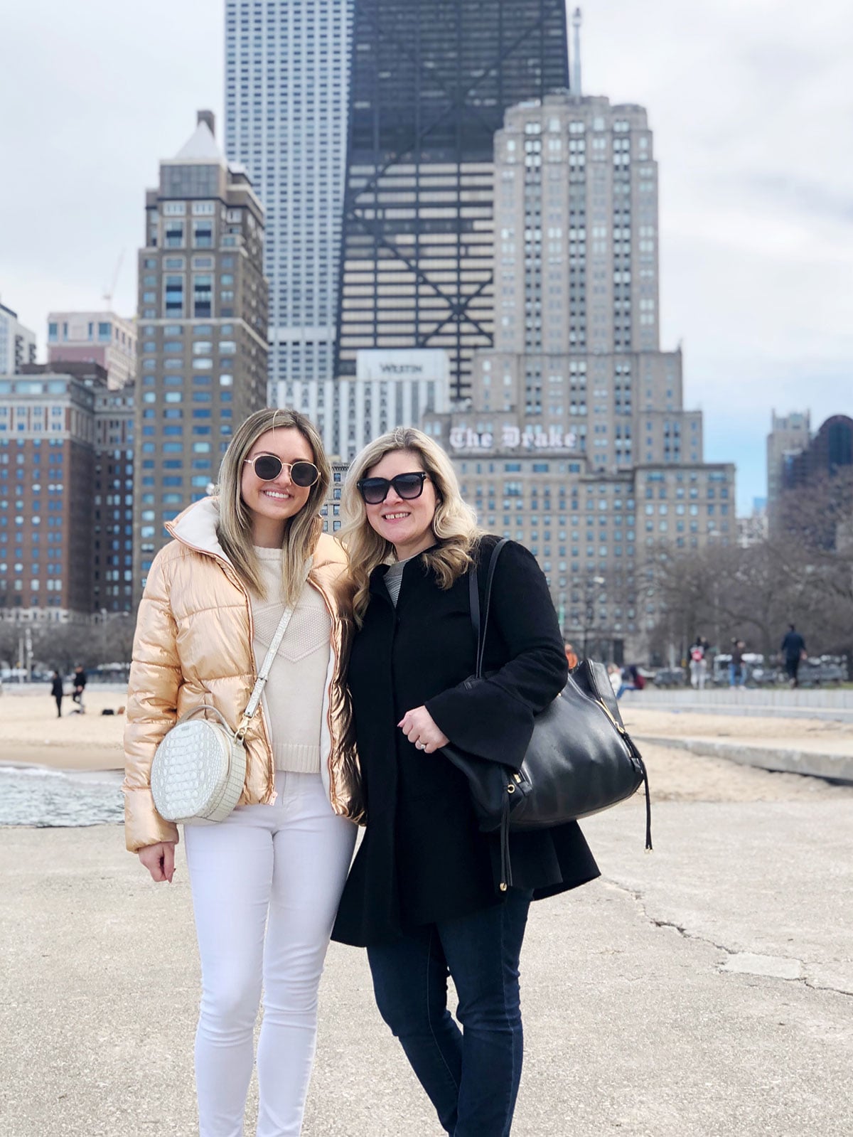 Bows & Sequins in front of the Chicago skyline near Oak Street Beach wearing a metallic puffer jacket and white jeans.