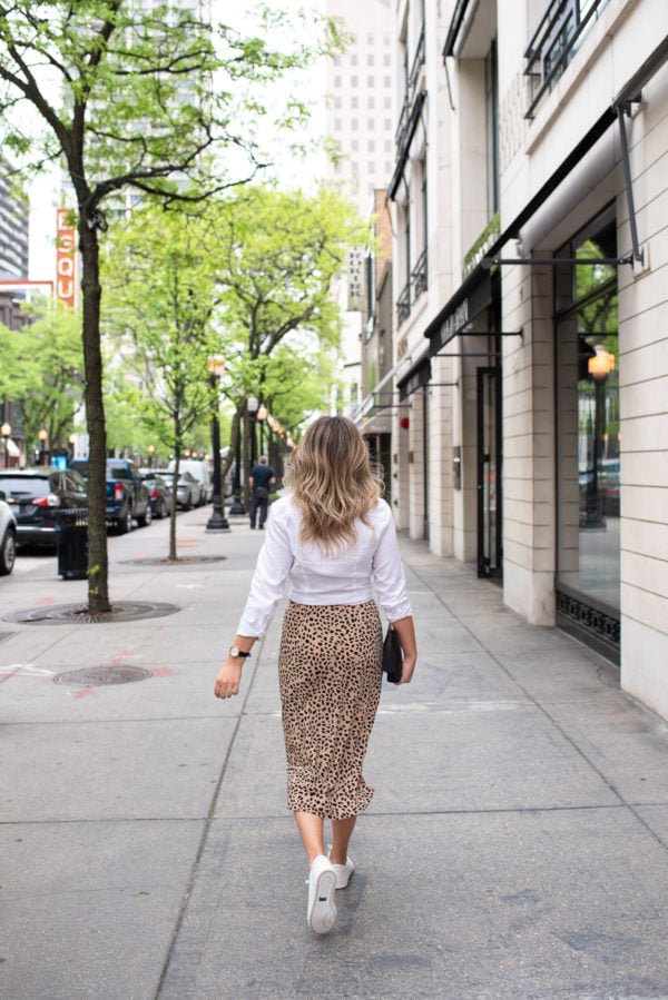 Fashion and lifestyle influencer Bows & Sequins styling a leopard midi skirt with a white button front shirt and sneakers.