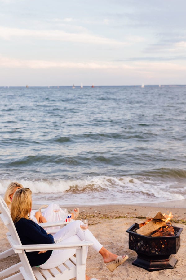 Two girls sitting in white adirondack chairs by a sunset beach bonfire at Wee Burn Beach Club in Connecticut