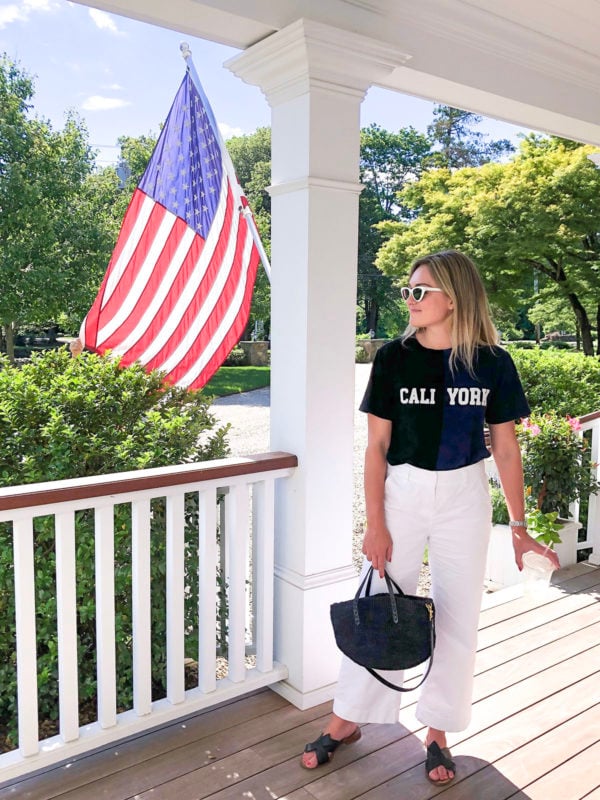 Lifestyle blogger Jessica Sturdy of Bows & Sequins wearing a Cynthia Rowley Cali York tee shirt with Vineyard Vines cropped wide leg white chino pants, a black Clare V straw tote, and black slides.