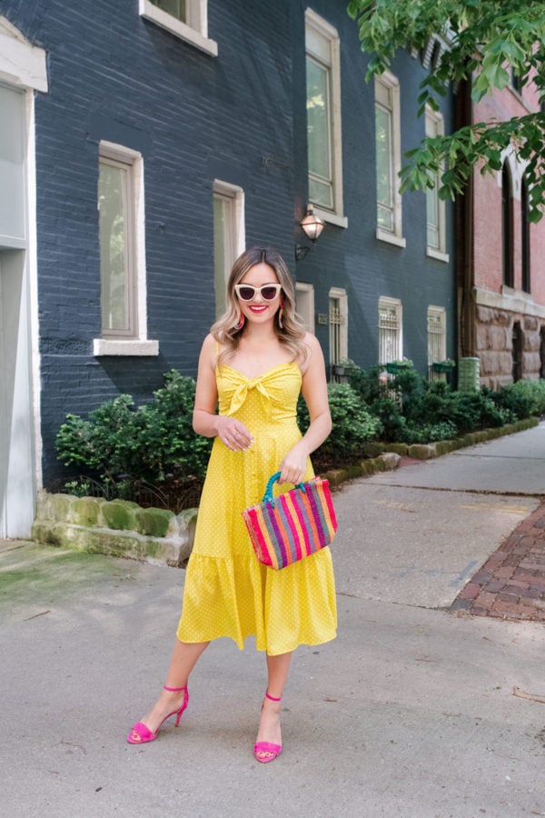 Chicago lifestyle and travel blogger Jessica Sturdy of Bows & Sequins styling a polka dot tiered midi dress with a colorful straw tote, Vince Camuto pink heels, and white sunglasses.