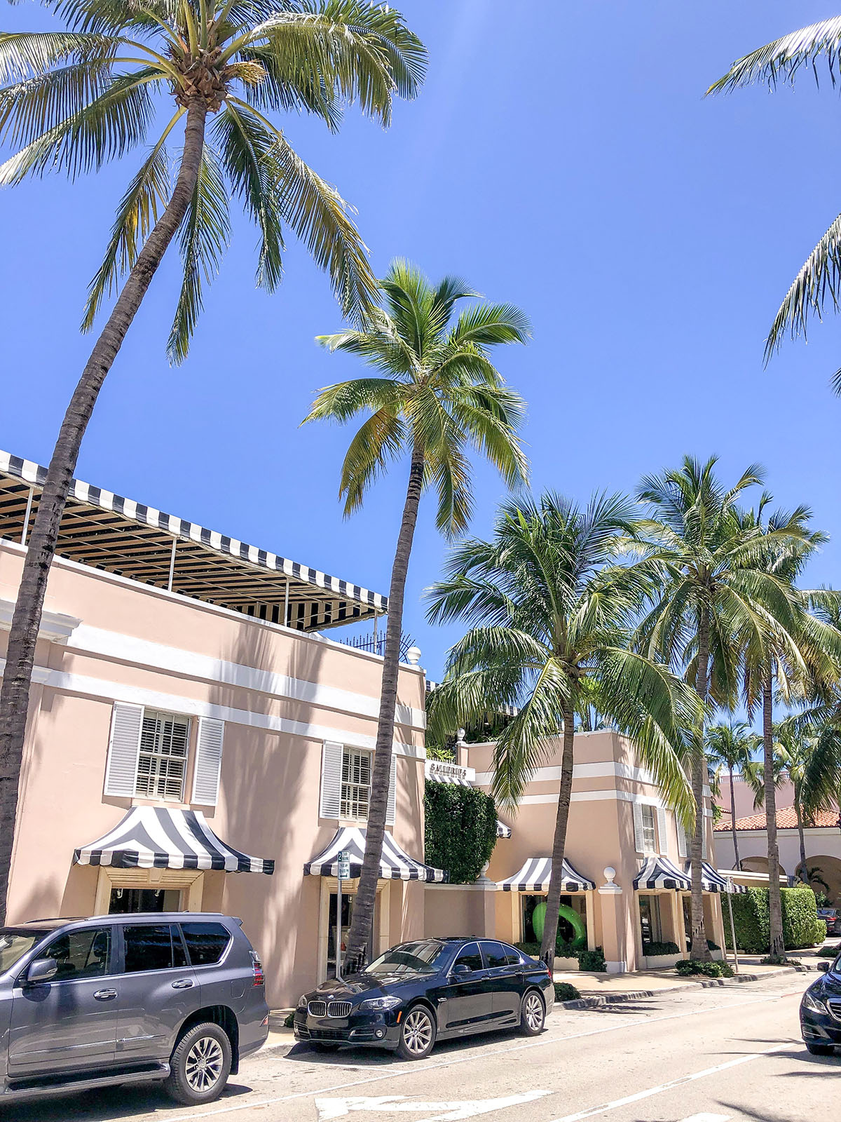 Pink building with black and white striped awnings in Palm Beach Florida with blue skies and palm trees on Worth Avenue.