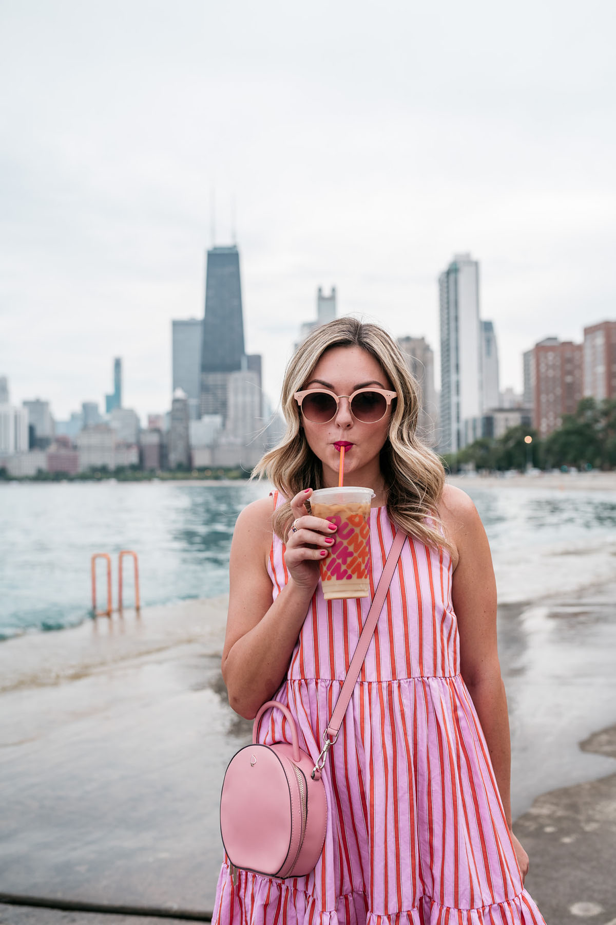Chicago blogger Jessica Sturdy of Bows & Sequins drinking a Dunkin' Donuts Iced Coffee in front of the Chicago skyline along the Lake Michigan lakefront path. She's wearing a pink and orange sleeveless, striped dress by MDS Stripes with a pink Kate Spade Andi Canteen Crossbody Bag.