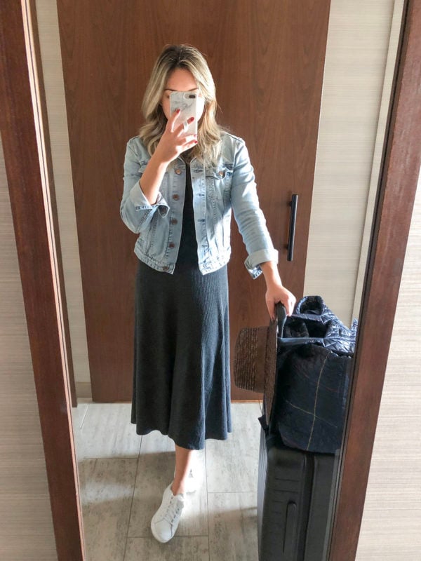 Travel blogger Bows & Sequins wearing a long ribbed sweater dress with a denim jacket and white sneakers with an Away suitcase and MZ Wallace travel bag.