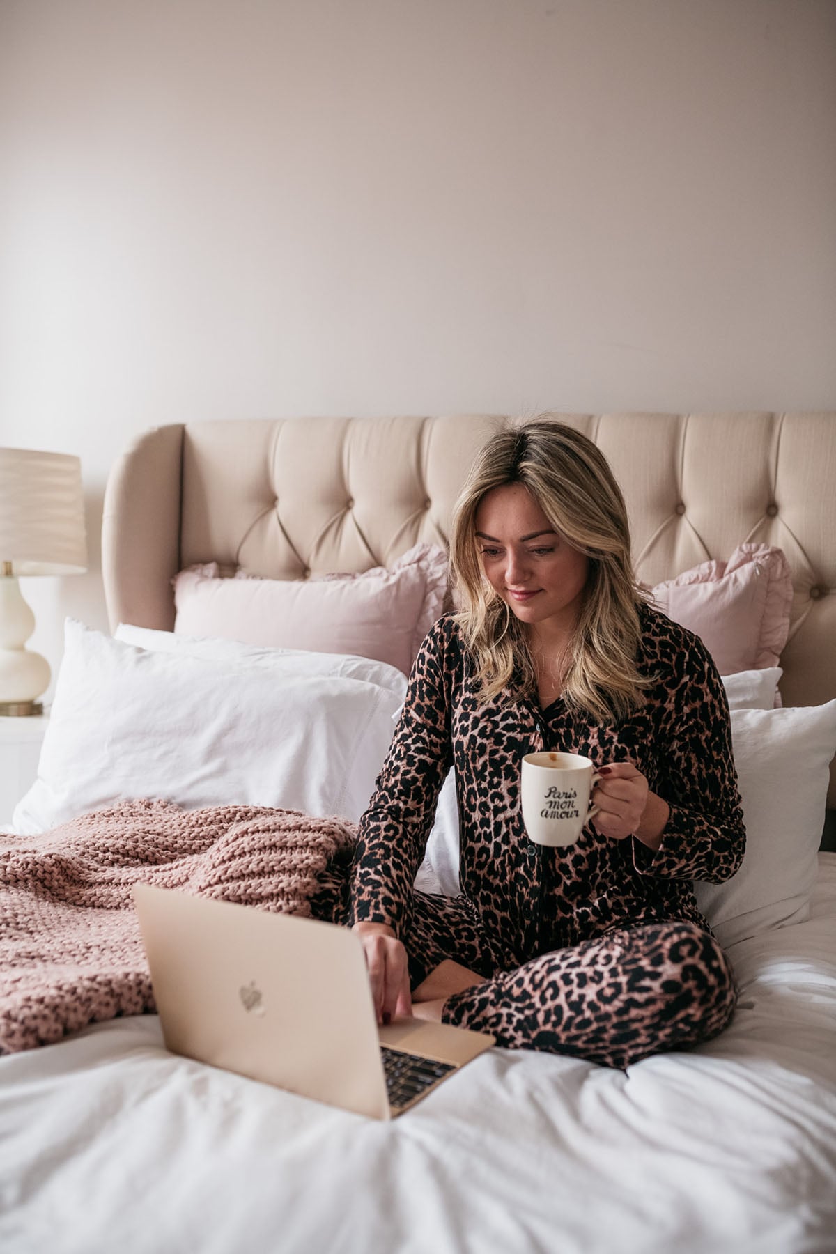 Chicago blogger Bows & Sequins wearing a leopard print pajama set in a cozy white bed and tufted headboard with a coffee mug and gold Macbook laptop.