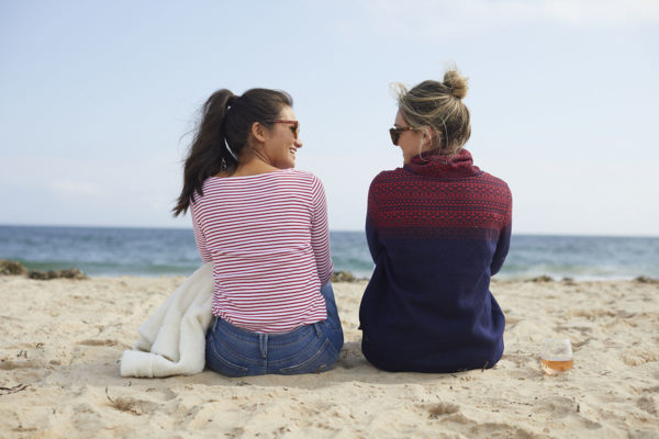 Bruntette in a red striped shirt and blonde in a blue and red fair isle funnelneck fleece on the beach.