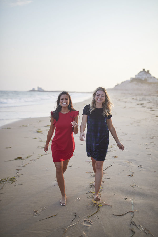 Caila Quinn wearing a red ruffled dress and Jessica Sturdy wearing a blackwatch plaid velvet dress on the beach at Ocean House Rhode Island with Taylor Swift's Watch Hill house in the background.