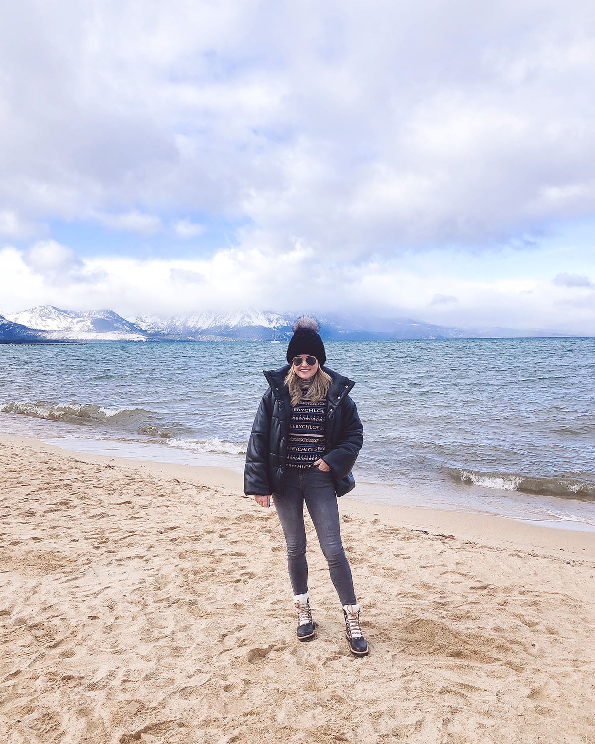 Travel blogger Jessica Sturdy of Bows & Sequins wearing a Nanushka Leather Puffer Coat, See by Chloe Logo Turtleneck, Grey Skinny Jeans, Black Fur Beanie, and Marc Fisher Shearling Booties by Lake Tahoe on the sand with snowy mountains in the back.