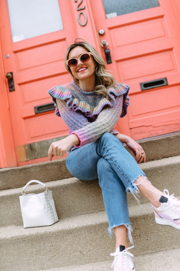 Chicago fashion blogger Jessica Sturdy that writes the blog Bows & Sequins wearing a rainbow ruffle sweater from LoveShackFancy with raw hem jeans and pastel sneakers in front of a coral door on Damen in Bucktown.