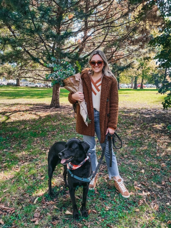 Jessica Sturdy and her black lab mix Marley at the Lincoln Park Green City Farmer's Market. She's wearing a brown teddy coat, varsity sweater, casual jeans, and high-top sneakers.