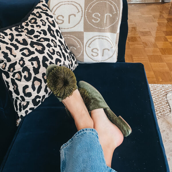 Home decor blogger Jessica Sturdy of Bows & Sequins wearing Birdies Pom Pom slippers on her navy velvet couch with a CB2 leopard pillow cover and custom BaubleBar initials blanket inspired by Hermes.