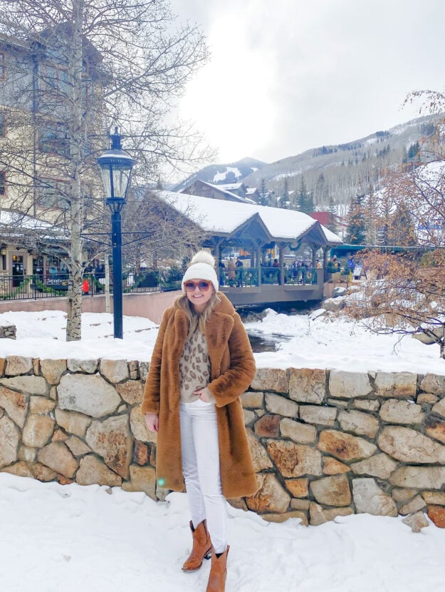 Fashion blogger wearing an Apparis Faux Fur Coat, leopard sweater, white velvet jeans, and cognac cowboy boots for a cute apres ski outfit in a mountain town.