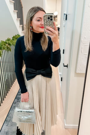 Lifestyle blogger Bows & Sequins wearing a fitted black turtleneck, cream pleated skirt, and a very oversized bow belt with a red lip and martini earrings.