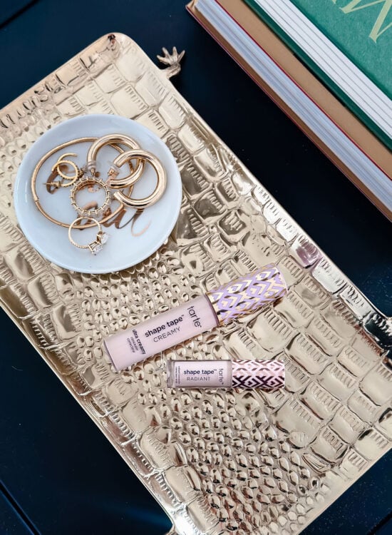 Tarte Shape Tape Concealer in a full-size tube of Ultra Creamy and a mini tube of Radiant on a brass crocodile tray with gold jewelry.