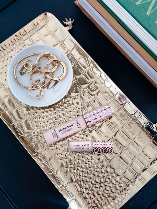 Tarte Shape Tape Concealer in a full-size tube of Ultra Creamy and a mini tube of Radiant on a brass crocodile tray with gold jewelry.
