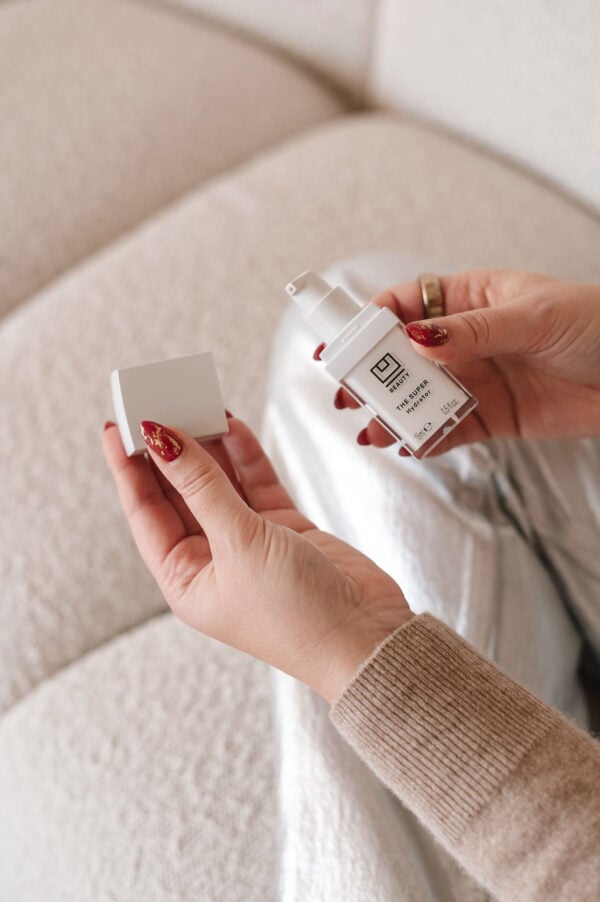 Chicago influencer holding U Beauty Super Hydrator wearing silver jeans and a tan cashmere sweater on a boucle couch.