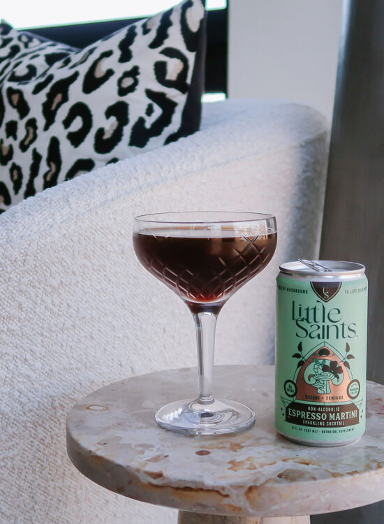 Little Saints Non Alcoholic Espresso Martini Canned Mocktail next to a crystal couple glass on a marble table with a boucle chair in the background.