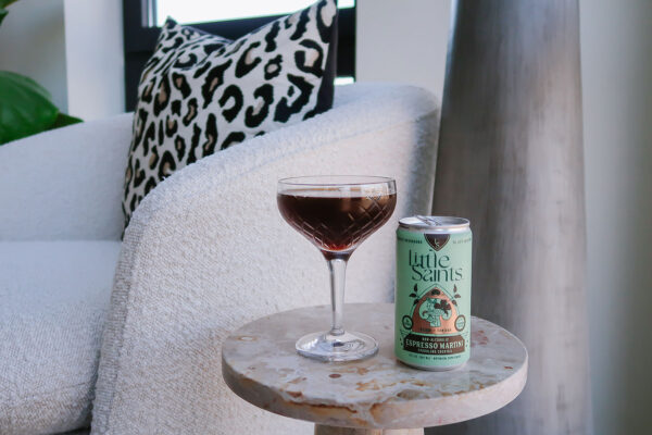 Little Saints Non Alcoholic Espresso Martini Canned Mocktail next to a crystal couple glass on a marble table with a boucle chair in the background.