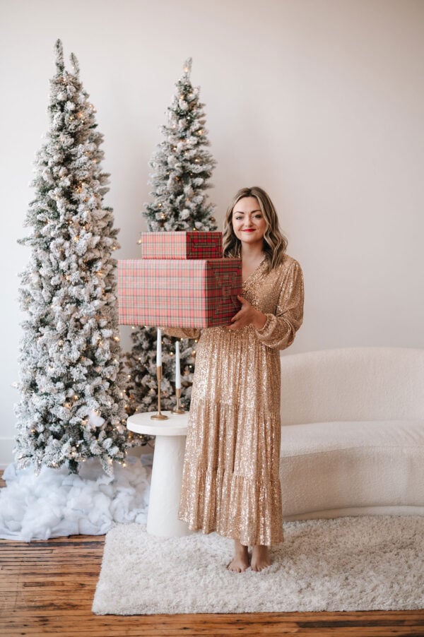 Fashion blogger Jessica Sturdy wearing a gold sequin maxi dress in front of white flocked Christmas trees and a boucle couch holding a stack of presents wrapped in tartan plaid paper.