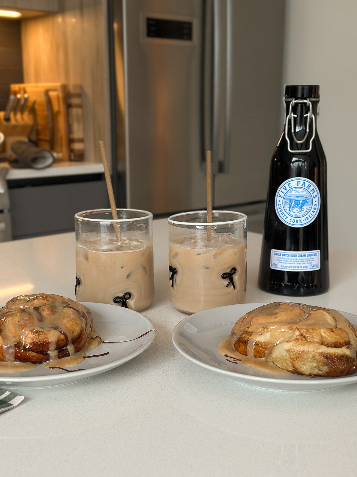 Two frosted cinnamon rolls on plates with iced coffee on a kitchen counter